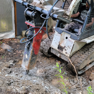 A tracked skid steer with a hydraulic breaker attachment breaking rocky ground near a structure
